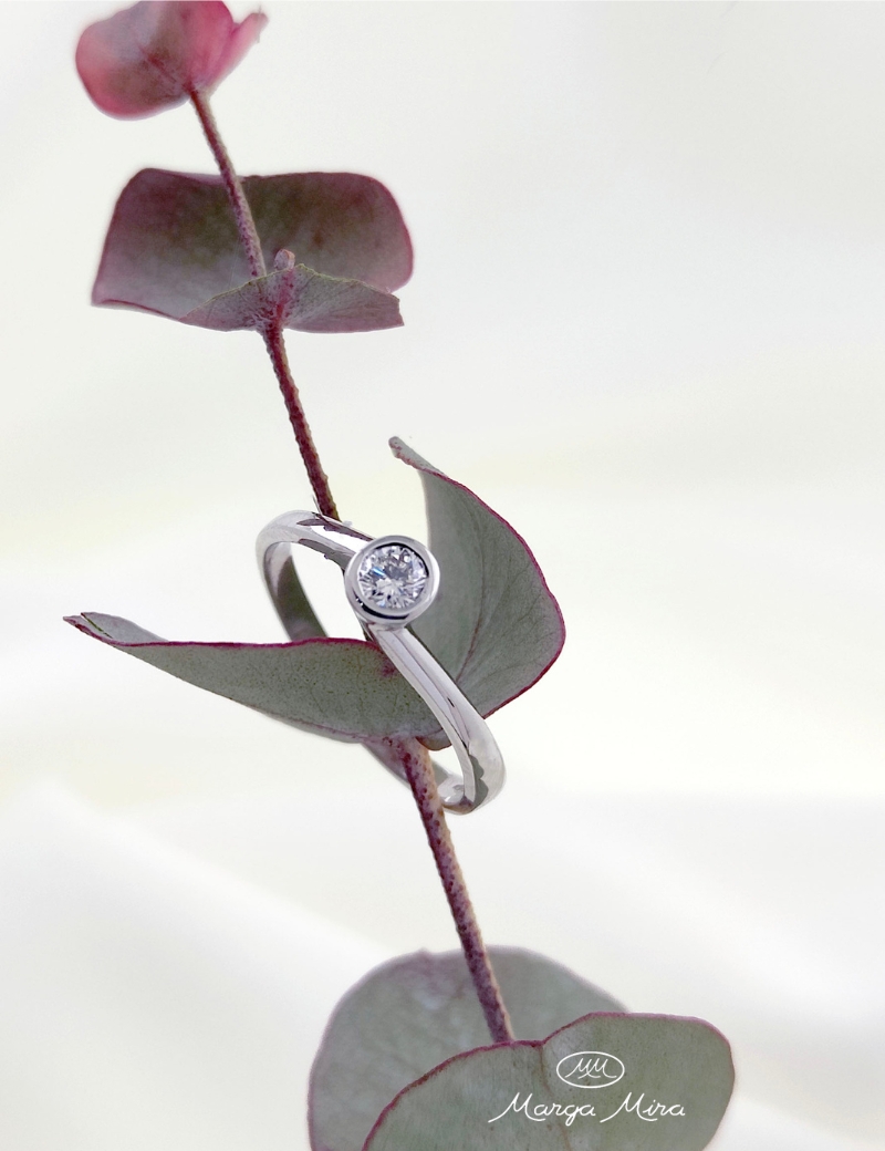anillo de diamante con engaste chaton o bisel sobre una tela de color blanco y rama de eucalipto